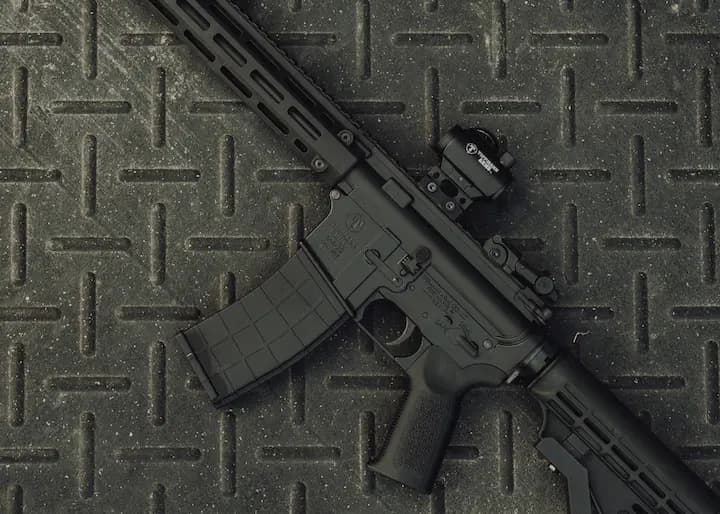 Close-up of a Tippmann Arms rifle featuring a mounted optic and textured magazine, resting on a rugged metal surface with a diamond plate pattern, showcasing durability and precision engineering.